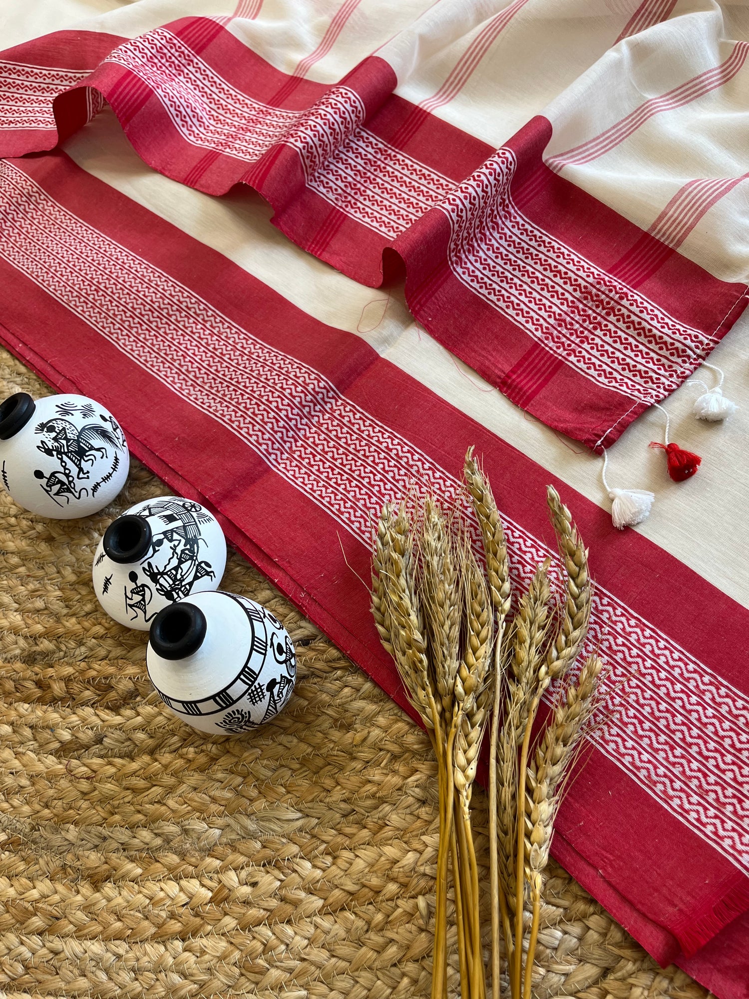 Traditional red and white saree with intricate woven patterns, displayed on a textured surface with decorative ceramic pieces and wheat stalks, showcasing the elegance of ethnic design.