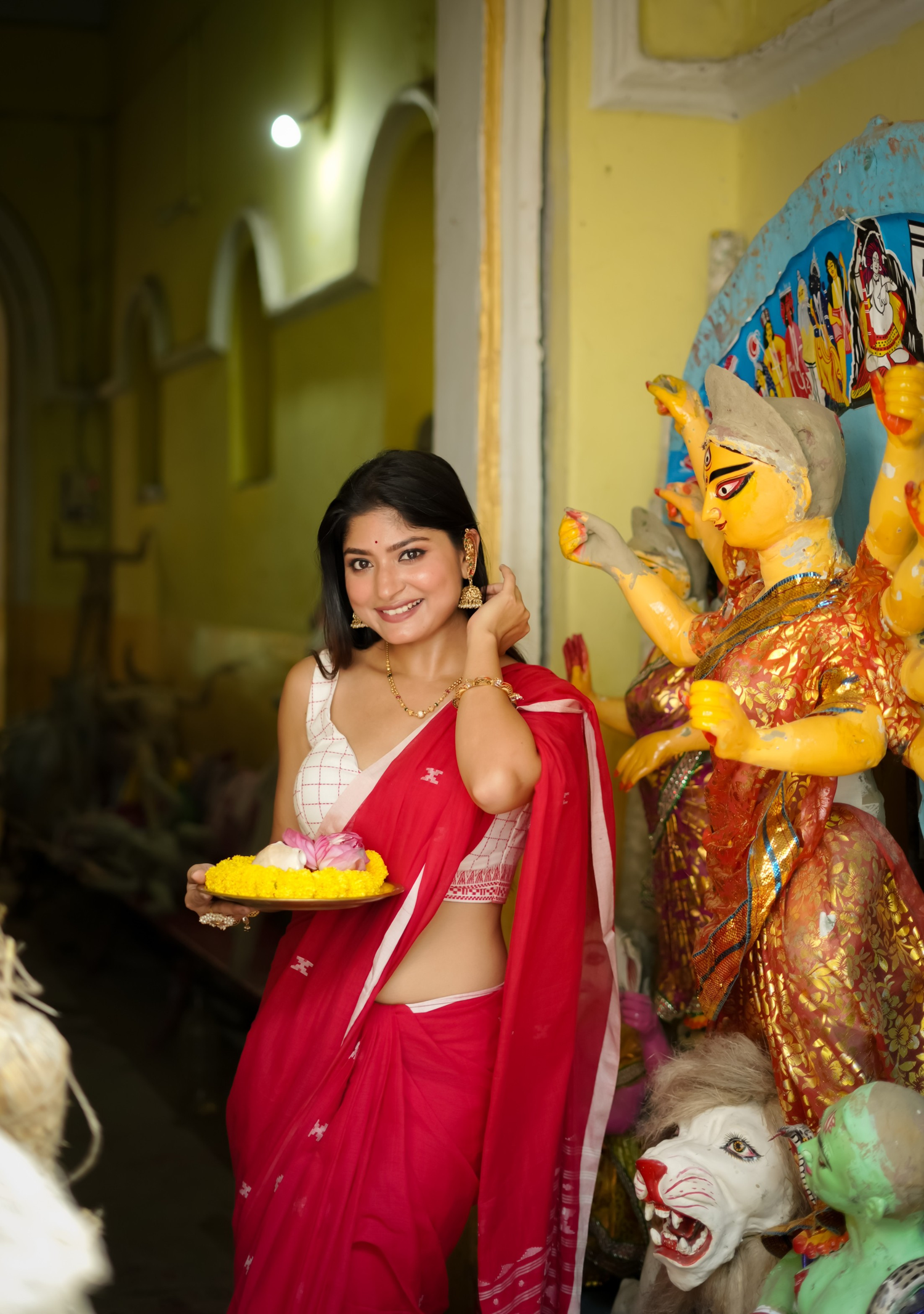 Smiling woman in a red saree with a white blouse holds a plate of flowers, standing beside a Durga idol. The scene reflects Durga Puja festivities, with vibrant decorations and traditional gold jewellery enhancing the festive atmosphere.